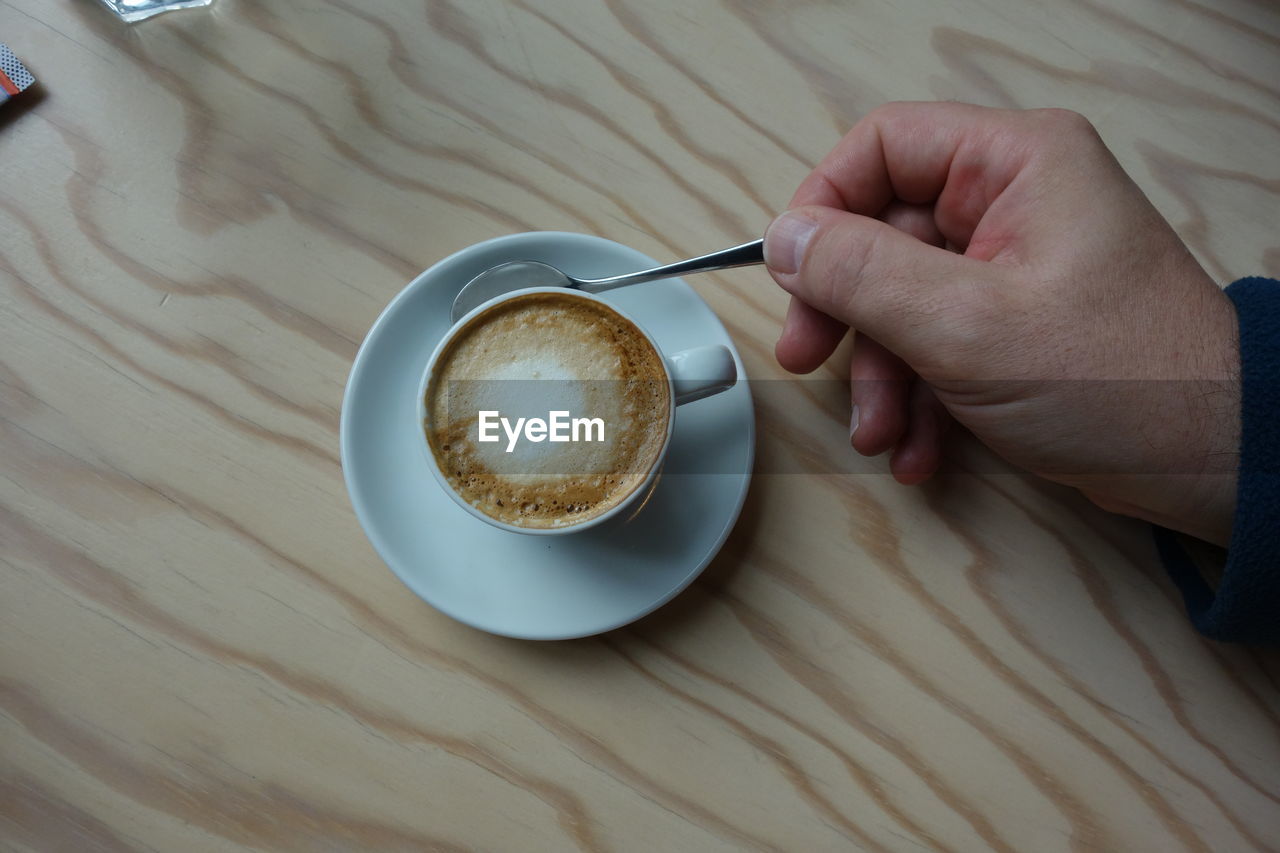 hand, coffee, food and drink, drink, mug, cup, coffee cup, one person, refreshment, table, adult, indoors, high angle view, hot drink, crockery, spoon, lifestyles, eating utensil, holding, kitchen utensil, saucer, food, cafe, wood, leisure activity, cappuccino, close-up, freshness, relaxation