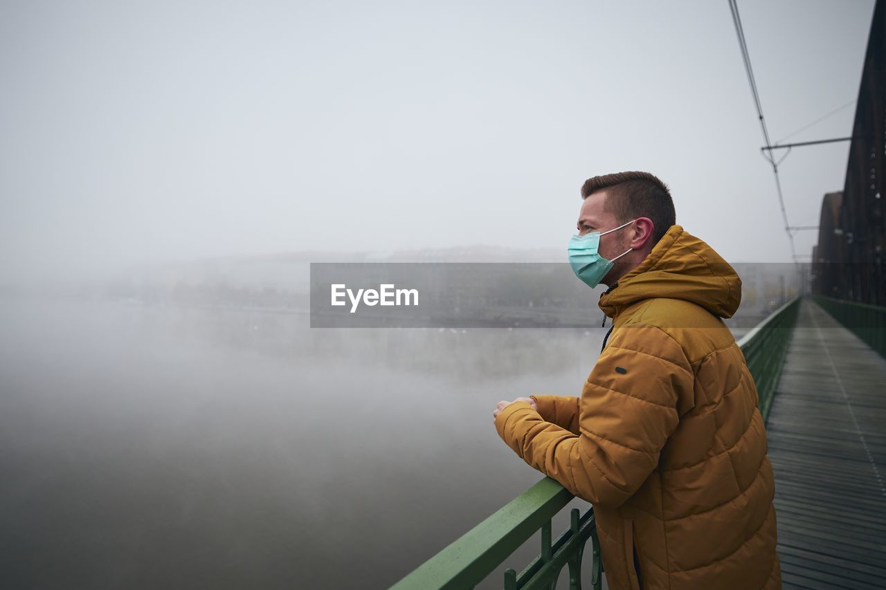 Side view of man standing by railing against lake during winter