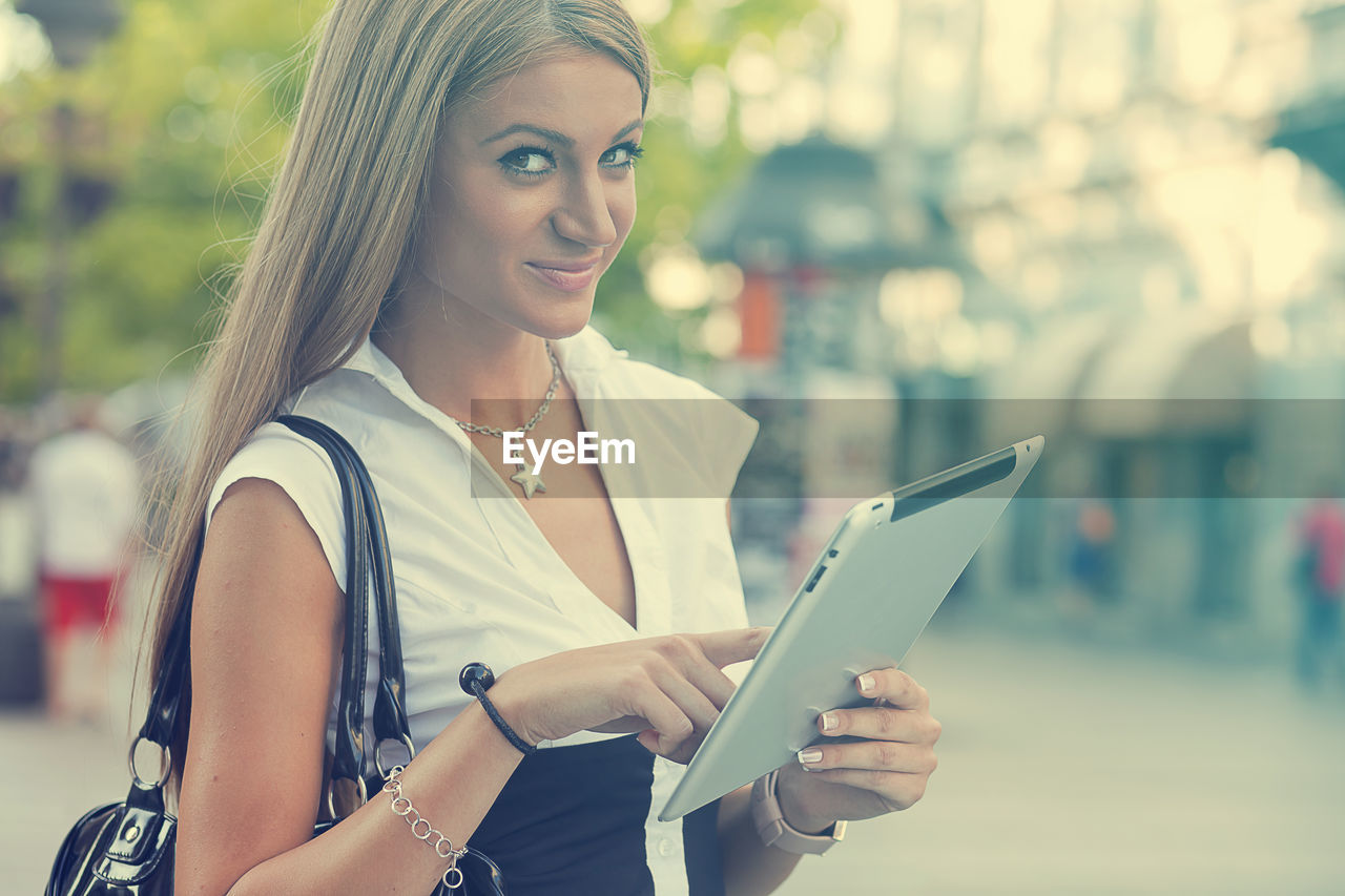 Portrait of young woman using digital tablet while standing on city street