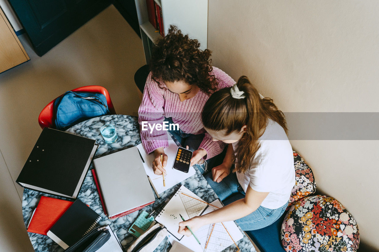 High angle view of young female friends using smart phone's calculator while homework