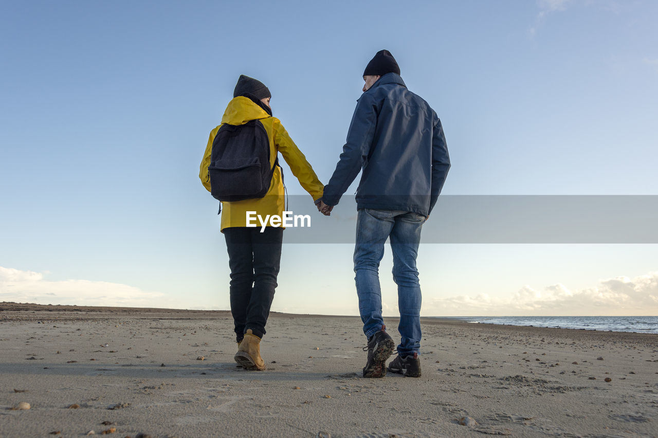 Rear view of a mature couple walking at beach holding hands, backpack and yellow coat, fall.