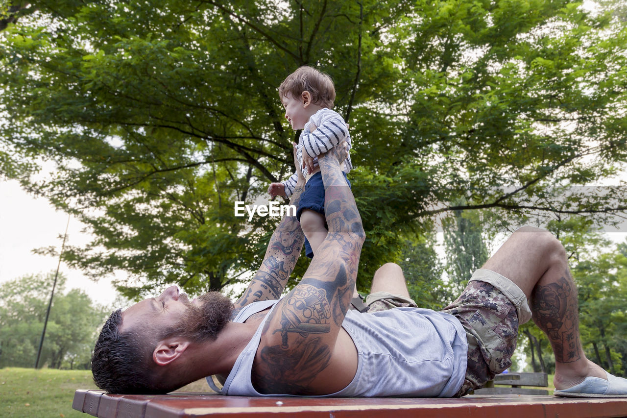 Father carrying son while lying on table against trees