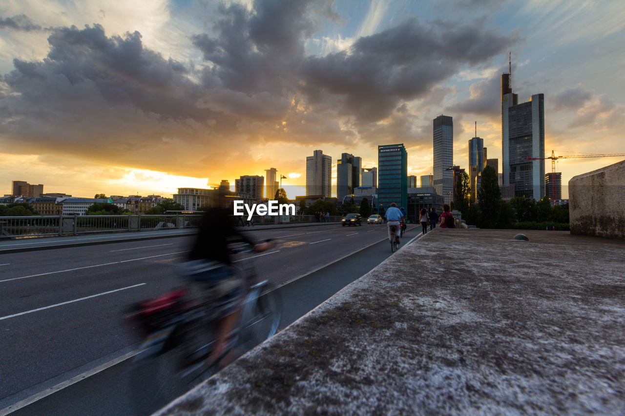 BLURRED MOTION OF PEOPLE ON ROAD BY BUILDINGS AGAINST SKY