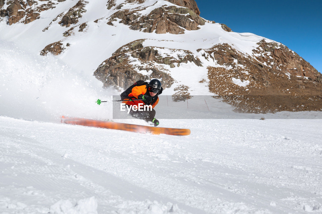 A skier in an orange jacket and a black helmet with a mask rides down a snowy slope at high speed
