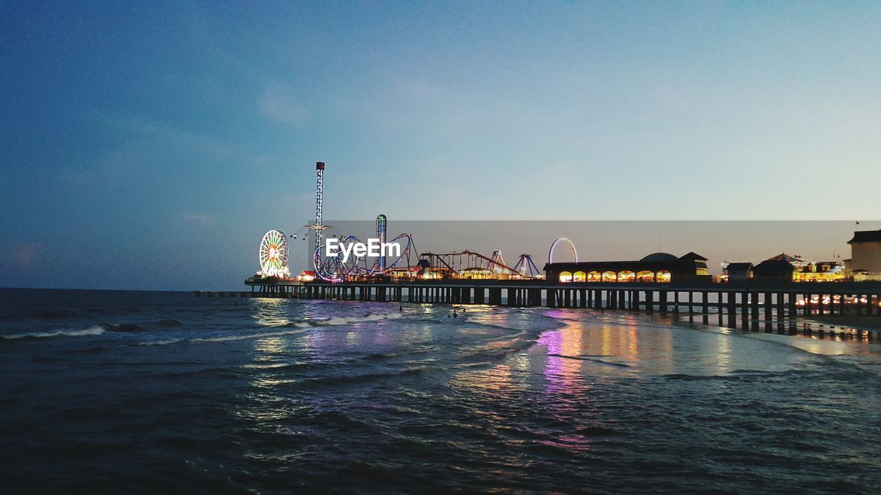 Illuminated amusement park on beach against sky at sunset