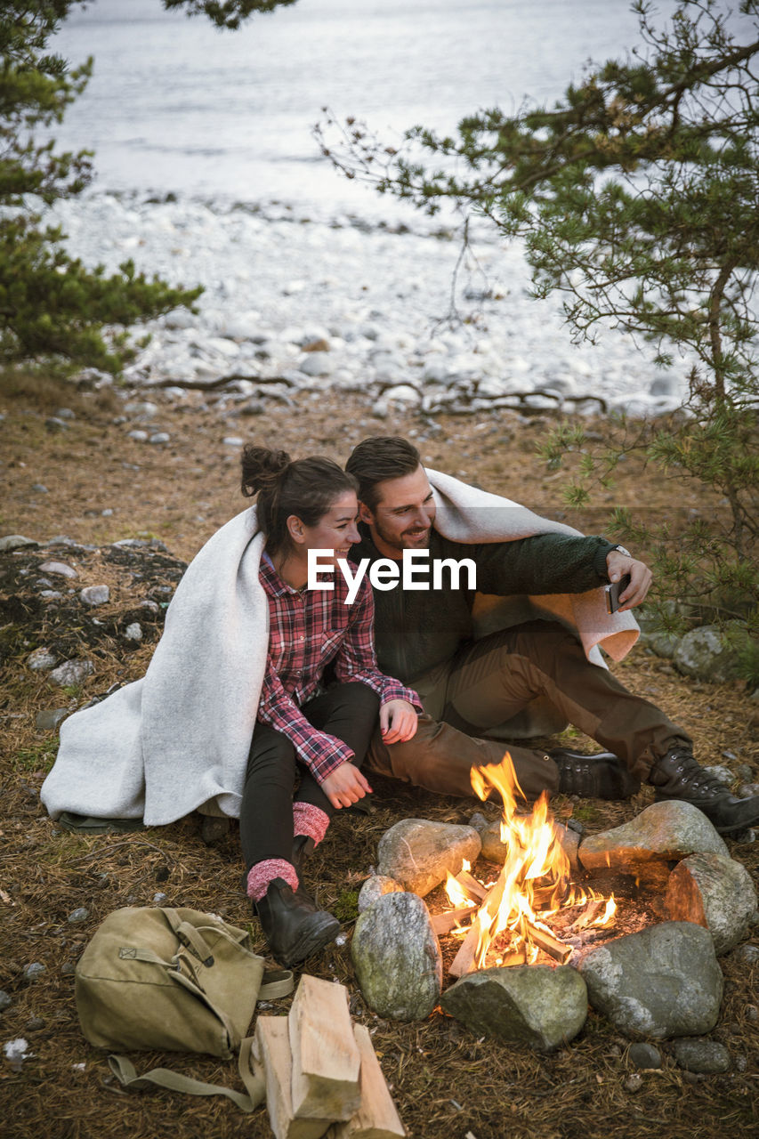 Couple wrapped in blanket taking selfie while sitting by fire pit at campsite