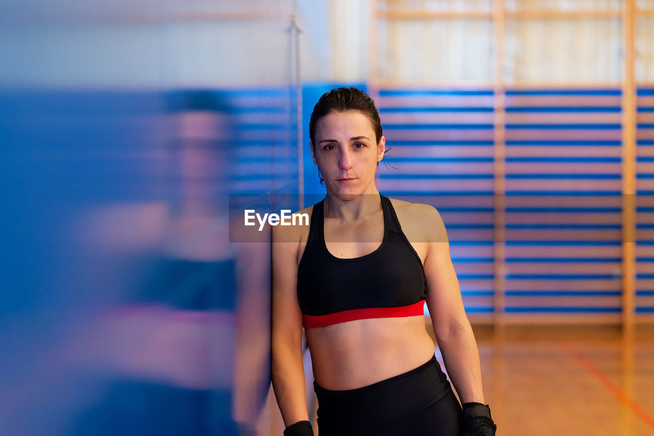 Female fighter with wraps leaning on blue wall at gym, looking camera