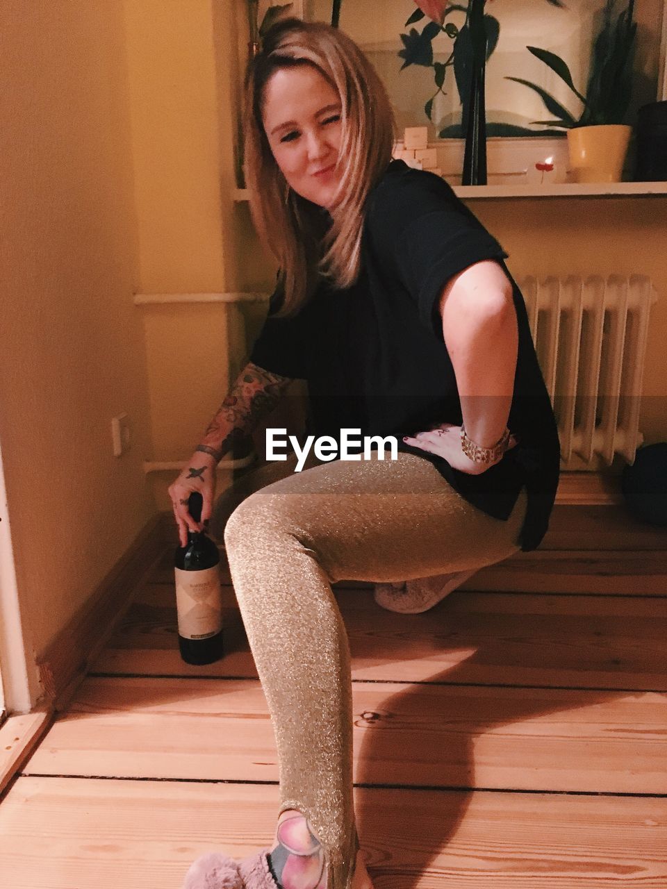 Portrait of happy woman holding wine bottle while crouching on hardwood floor