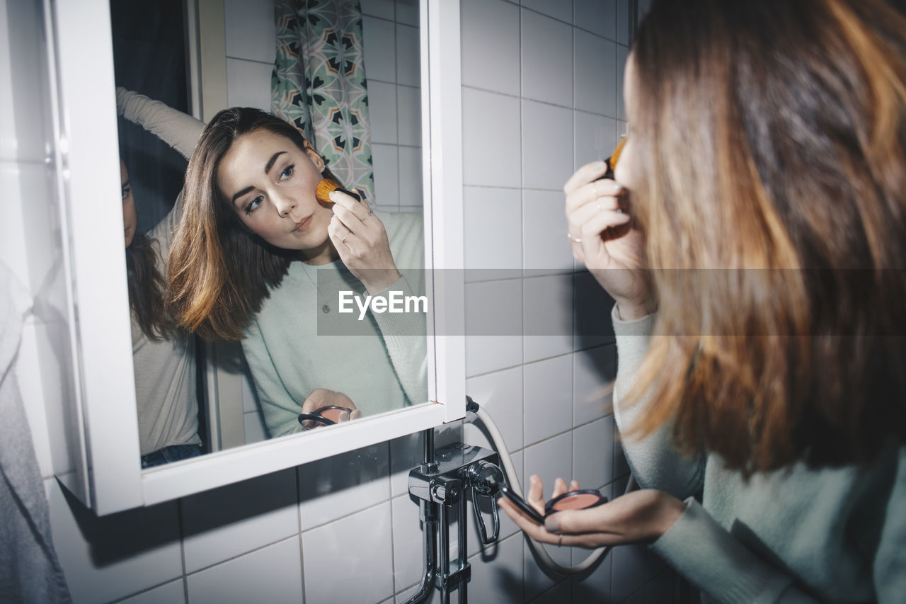 Young woman applying blush looking in mirror at college dorm bathroom