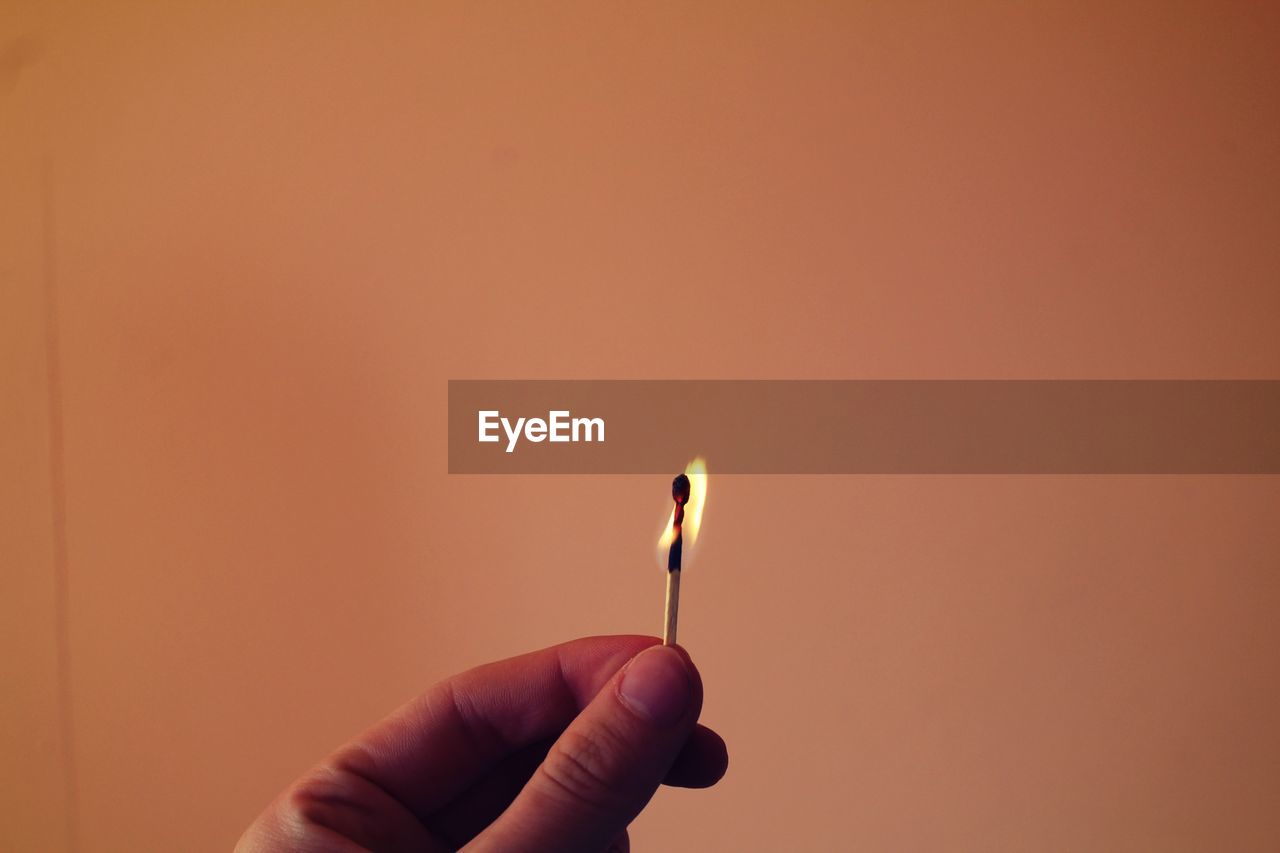 Cropped hand holding burning matchstick against brown wall