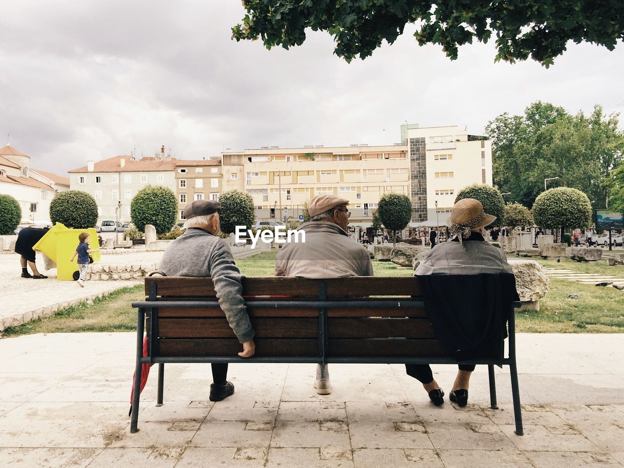 REAR VIEW OF PEOPLE SITTING ON BENCH AGAINST BUILDINGS