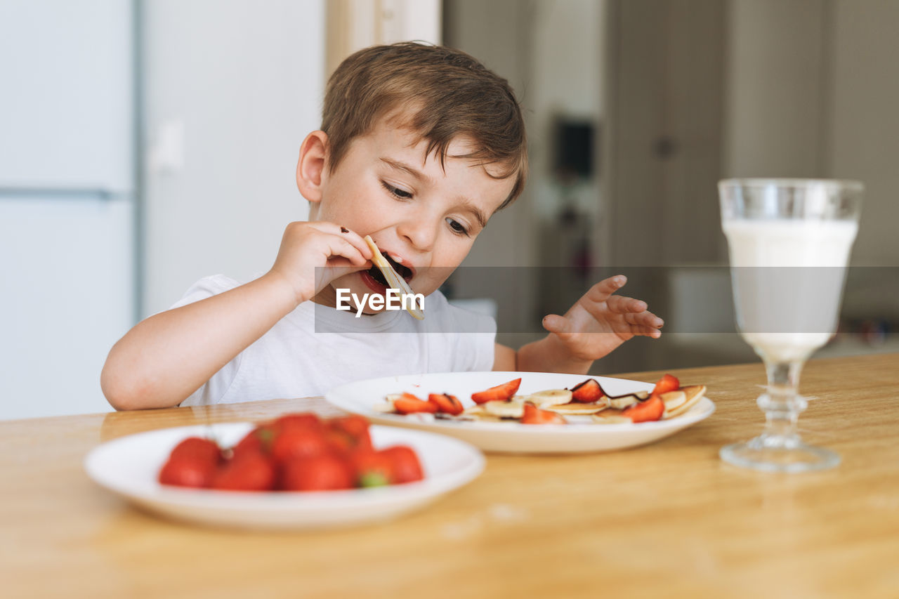 Cute toddler boy having breakfast with puncakes with berries and glass of milk in kitchen at home