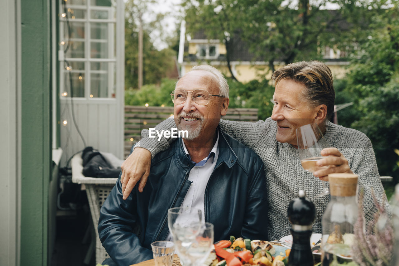 Smiling male friends sitting at dining table enjoying dinner party in back yard