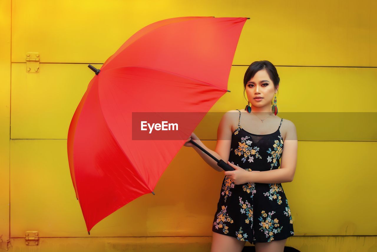 Portrait of young woman with red umbrella standing against yellow wall