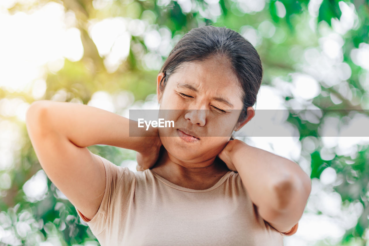 Woman with eyes closed touching neck in pain against trees