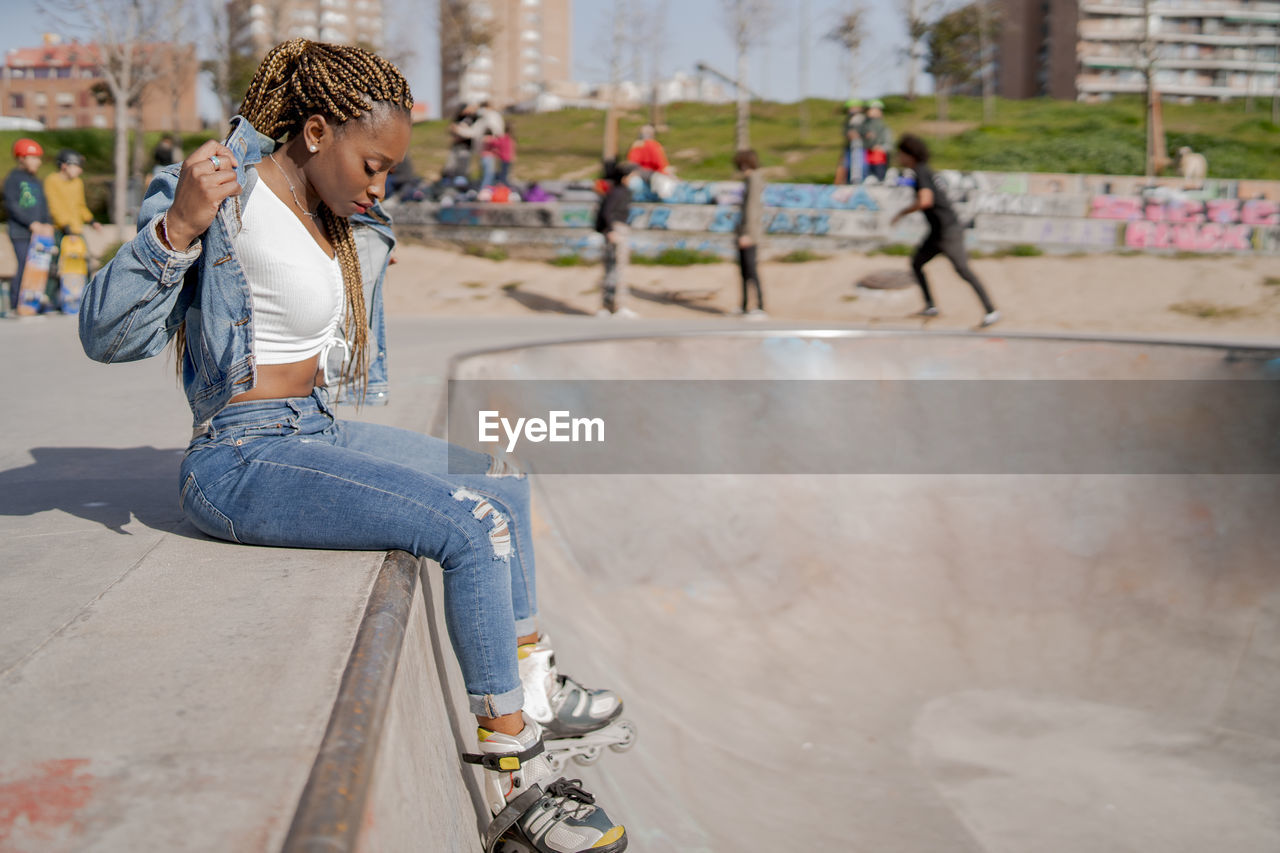 Cool black female with braided hairstyle and in rollerblades sitting on ramp in skate park and looking down