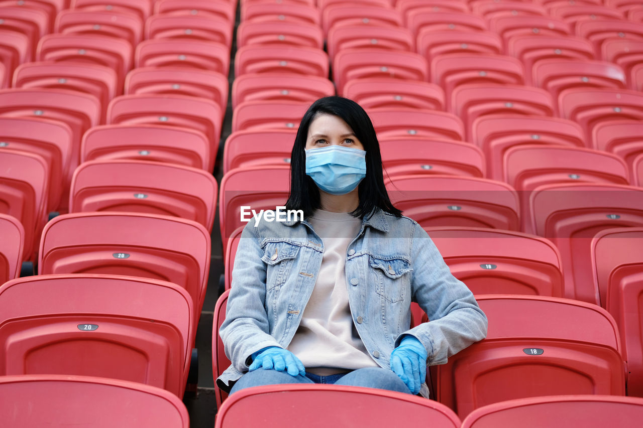 A female fan in a medical mask and rubber gloves sits alone in an empty stadium with red seats.