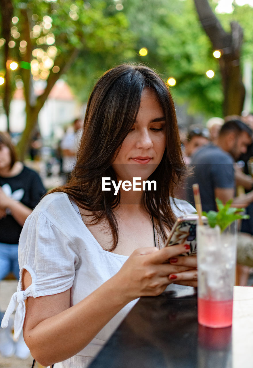 Young woman using mobile phone, drinking cocktail in open air bar in park in city