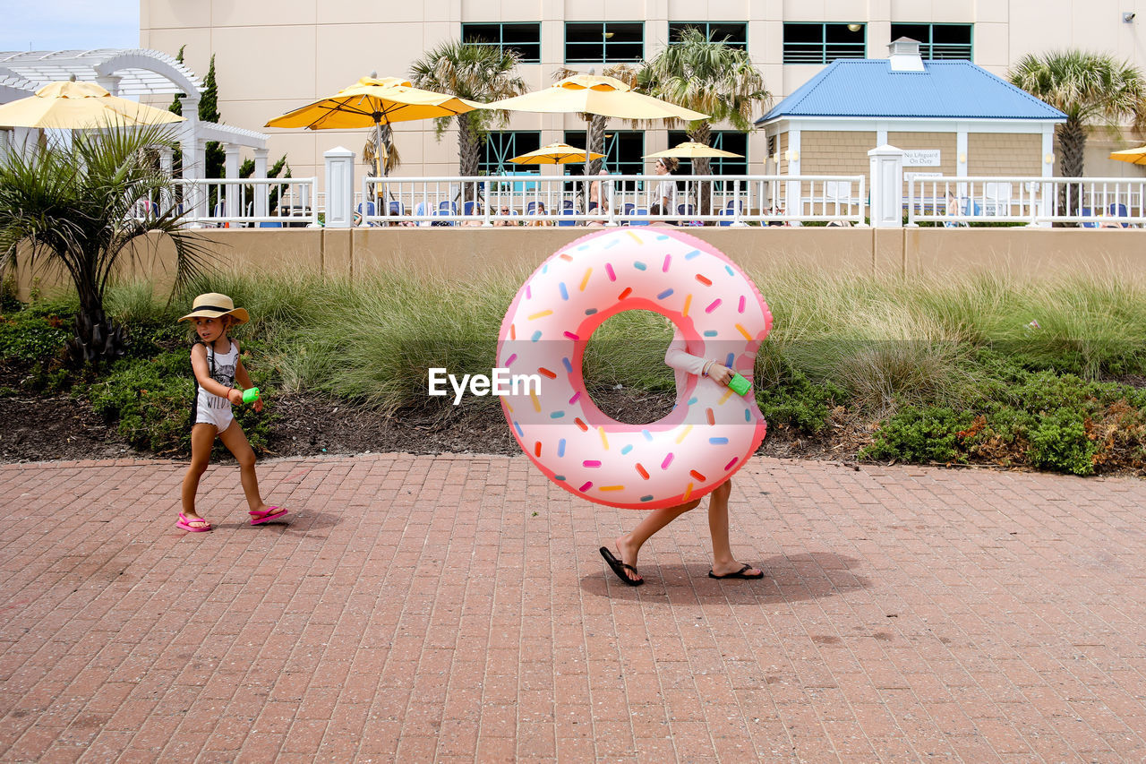 Two girls walking to pool with oversized donut raft wearing hat