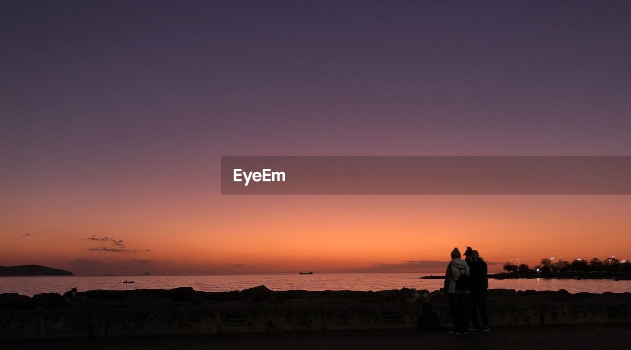 Male and female sitting by sea against sky during sunset