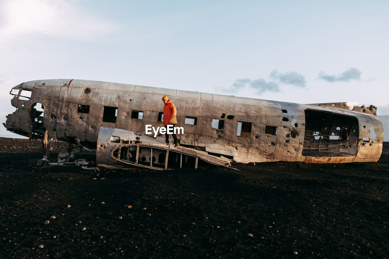 Side view of young tourist standing on wrecked aircraft between deserted lands and blue sky