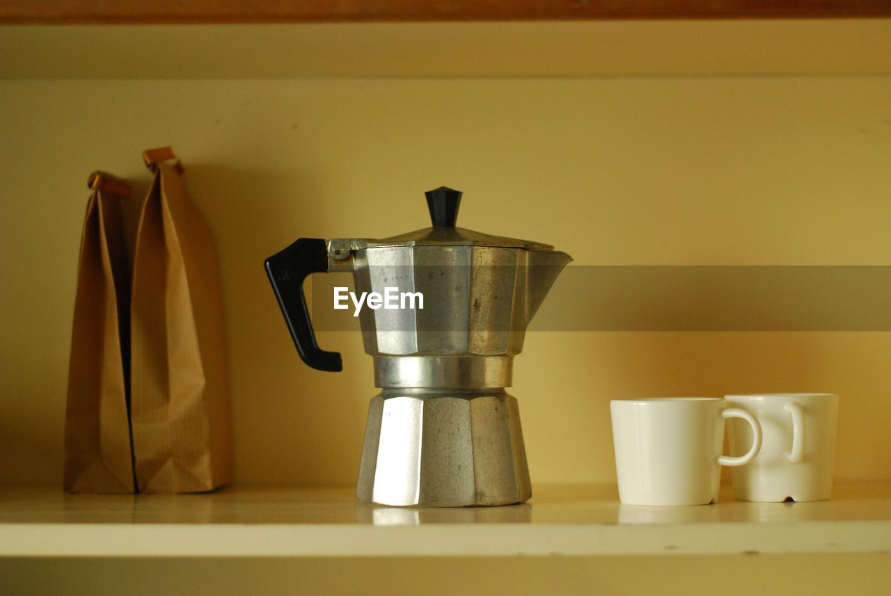 Close-up of coffee maker with cups and paper bags on shelf