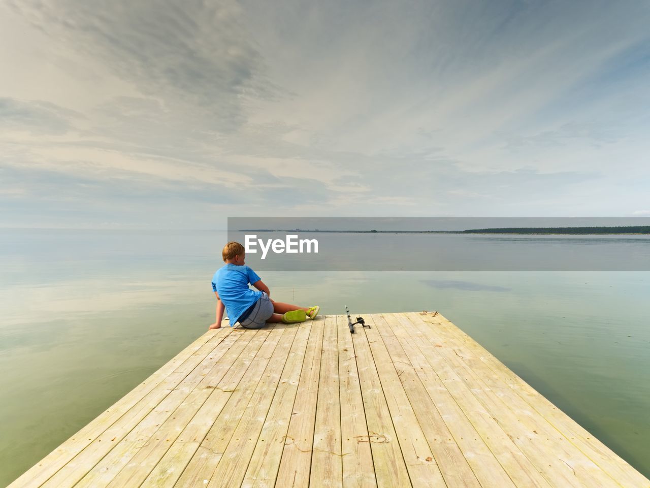 Small fisher on mole. blond boy in blue t-shirt, grey shorts and green flip-flops fishing on pier