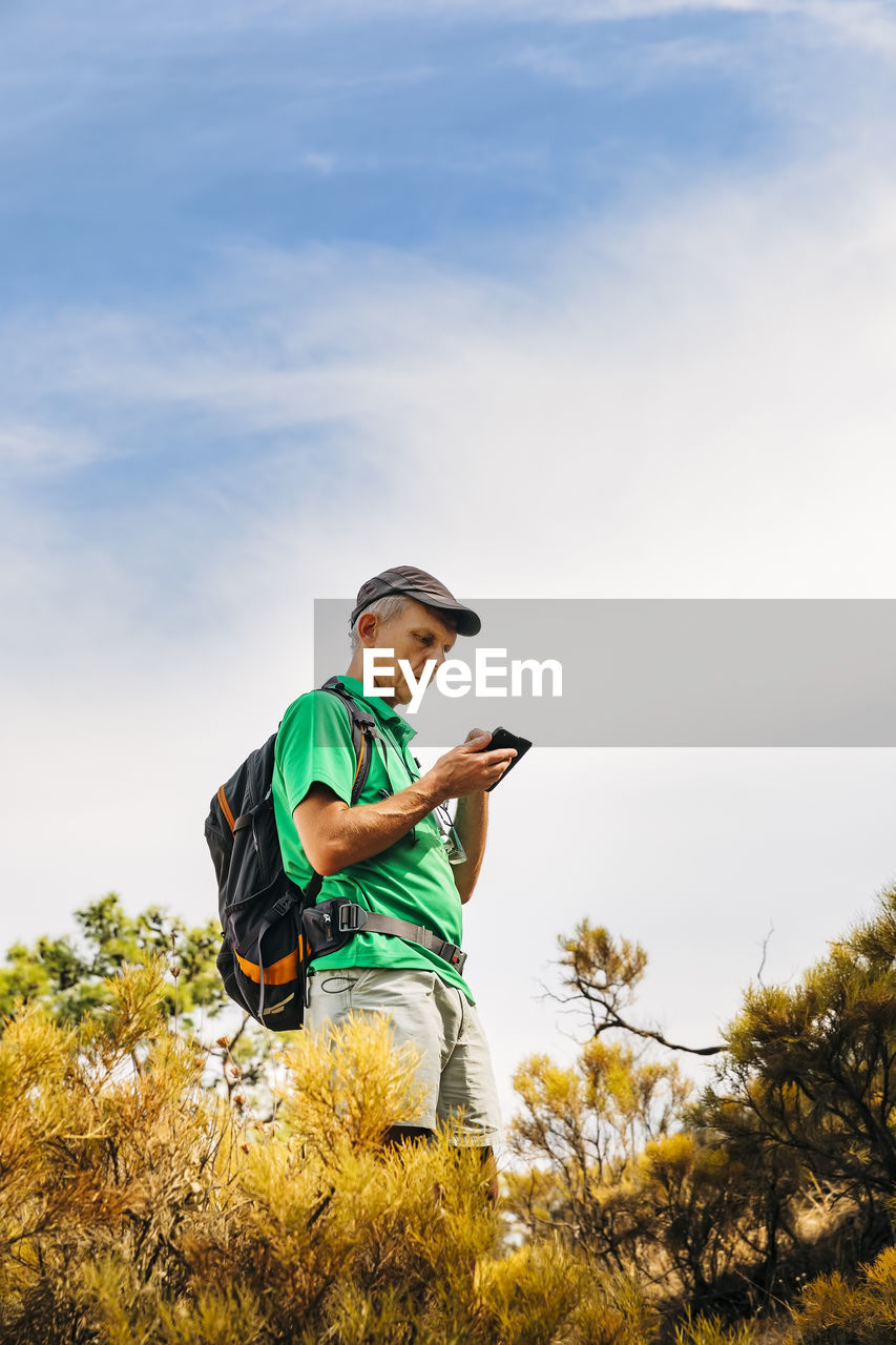 Senior backpacker using smart phone on cliff against sky