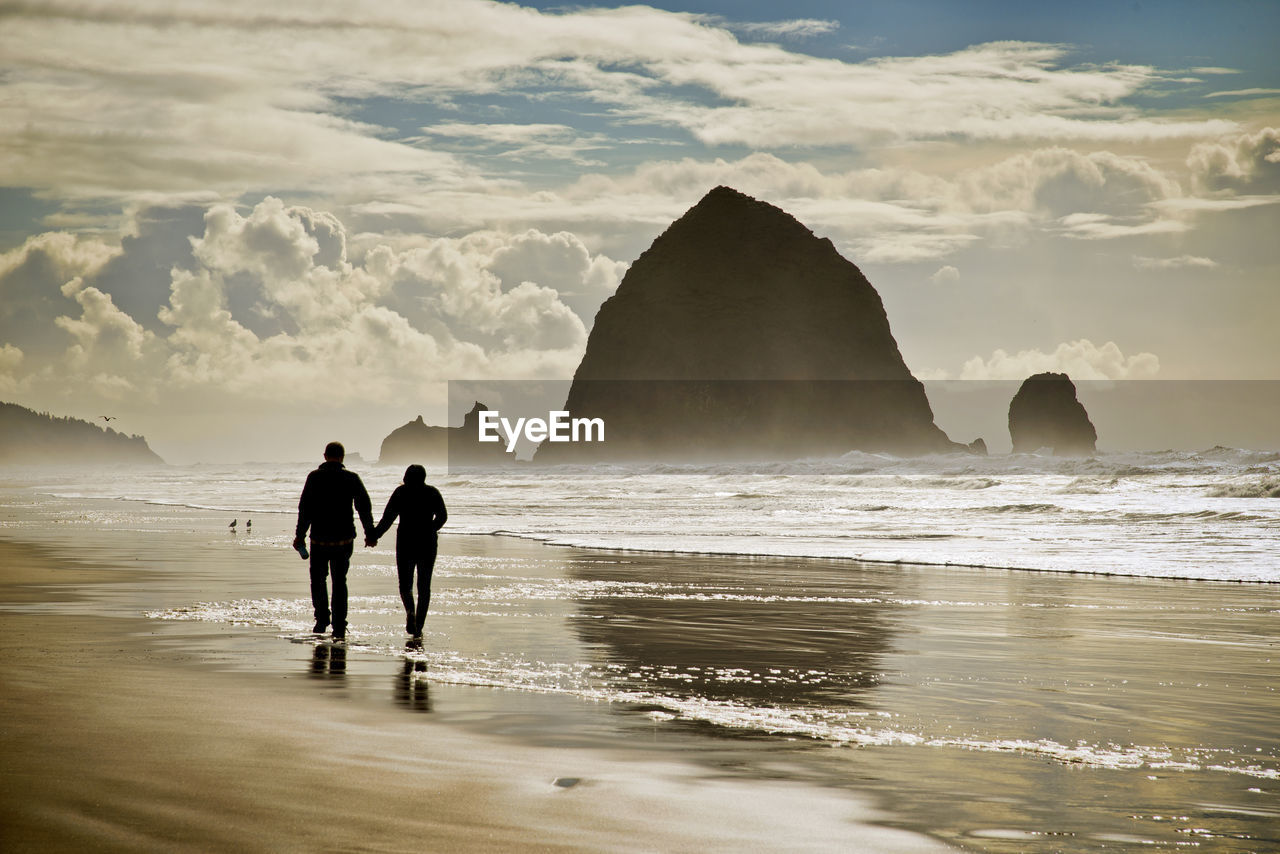 Silhouette man and woman walking at beach against cloudy sky