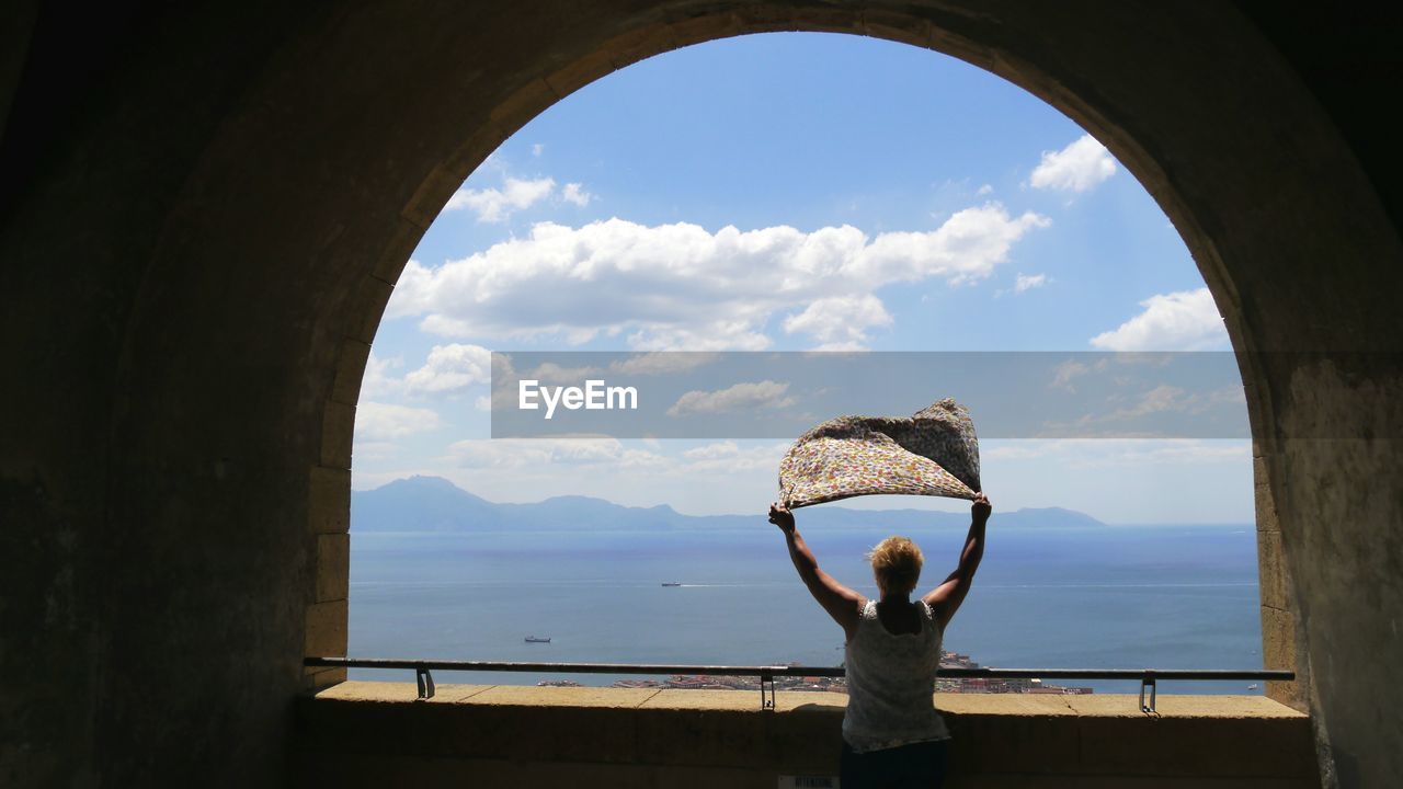 Rear view of woman holding scarf while standing in arch balcony against sky