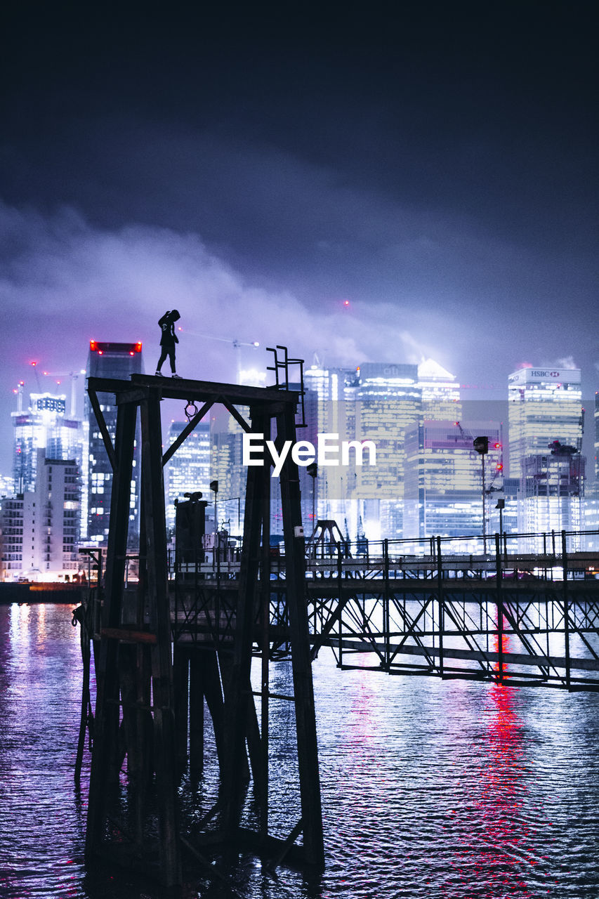 Silhouette man standing on pier over river against sky in city