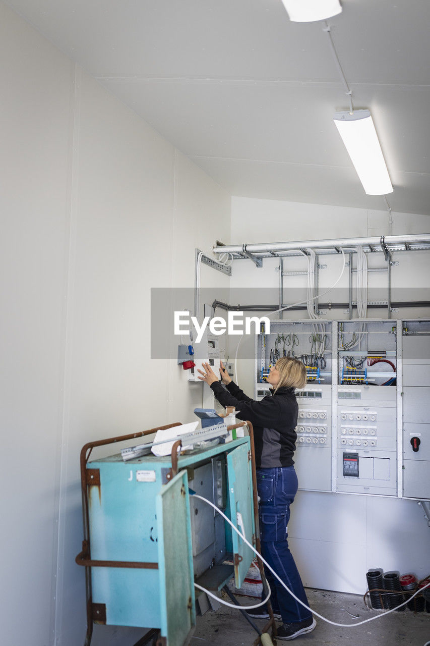 Female electrician installing fuse box while standing in control room at industry