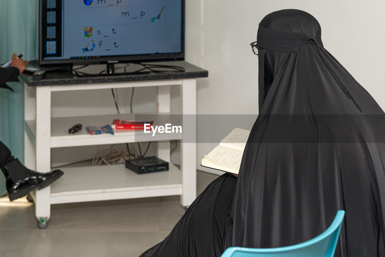 Muslim woman in  full hijab taking notes from the educational material on the screen