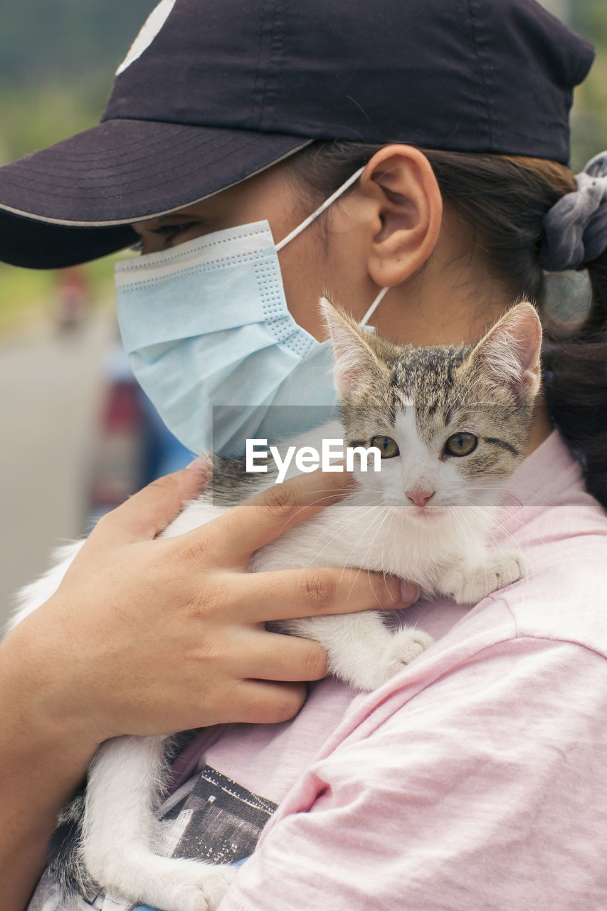 Unrecognizable woman wearing face mask and black cap carrying a young white and gray cat on her shoulders in a blurred natural environment