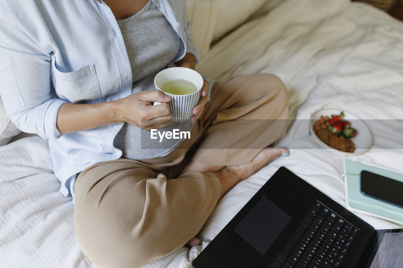 Pregnant businesswoman holding tea cup sitting with laptop on bed