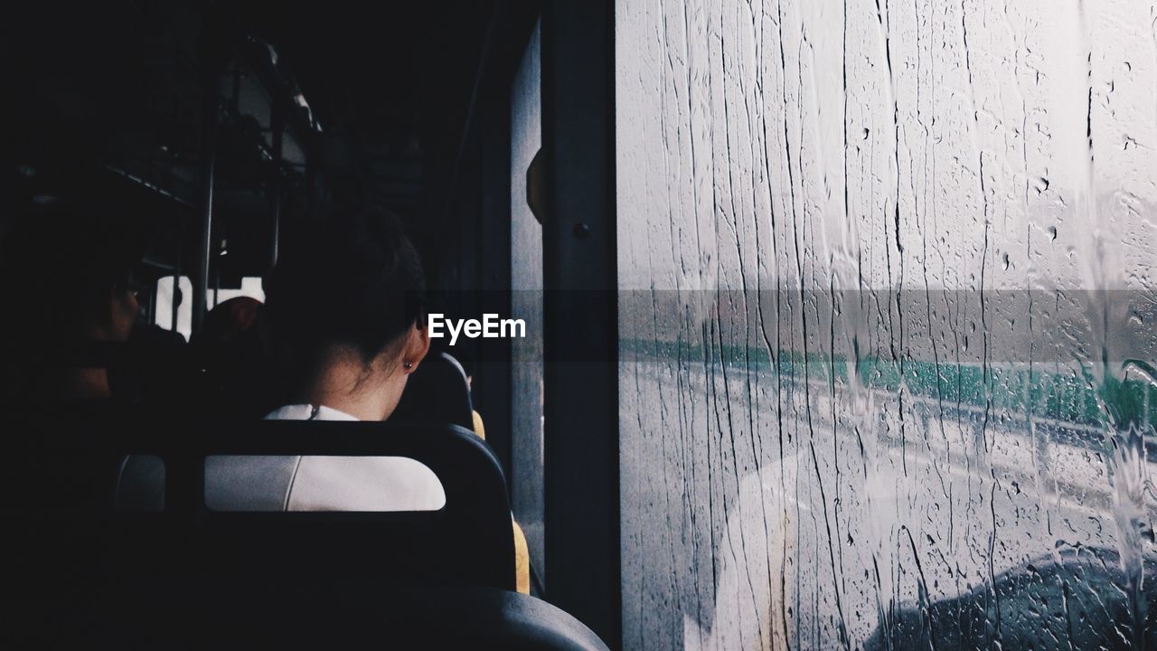 Rear view of woman relaxing by wet window in bus during rainy season