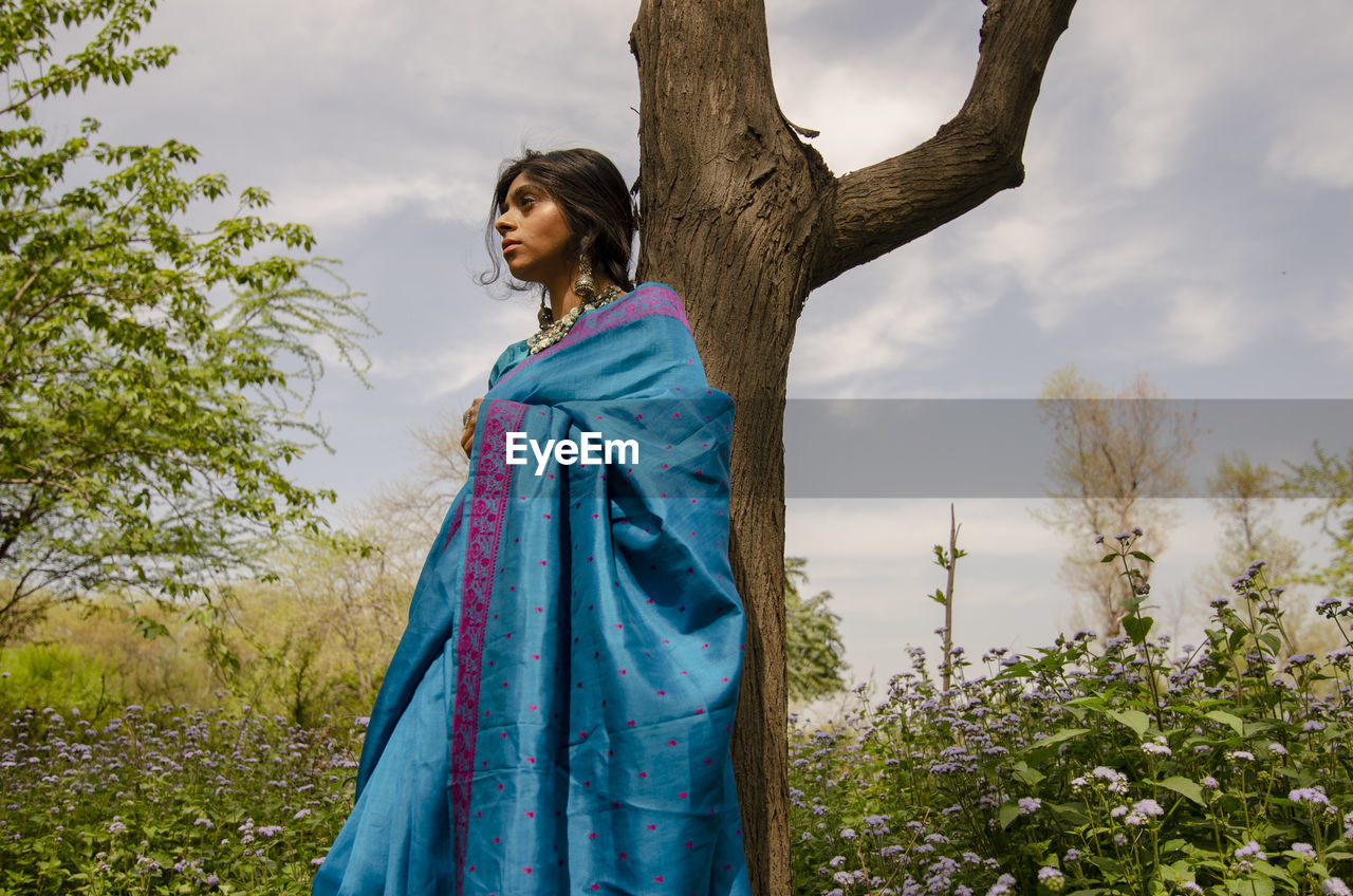 Young woman looking away while standing by tree against sky