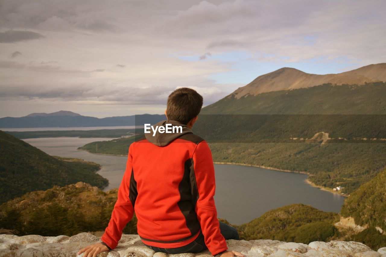 Rear view of man sitting on cliff lake and mountains against cloudy sky