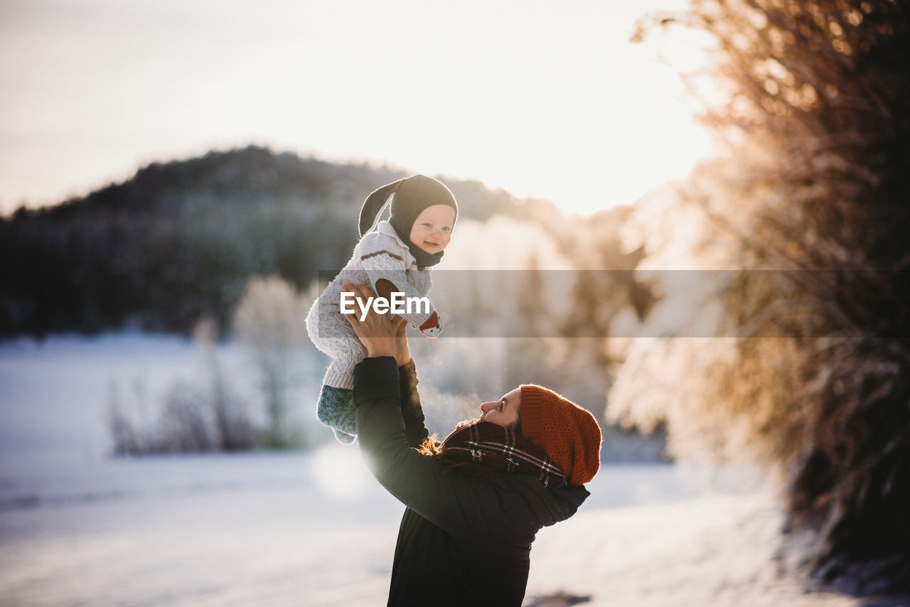 Mum holding smiling baby up in winter on sunny snowy day in the forest