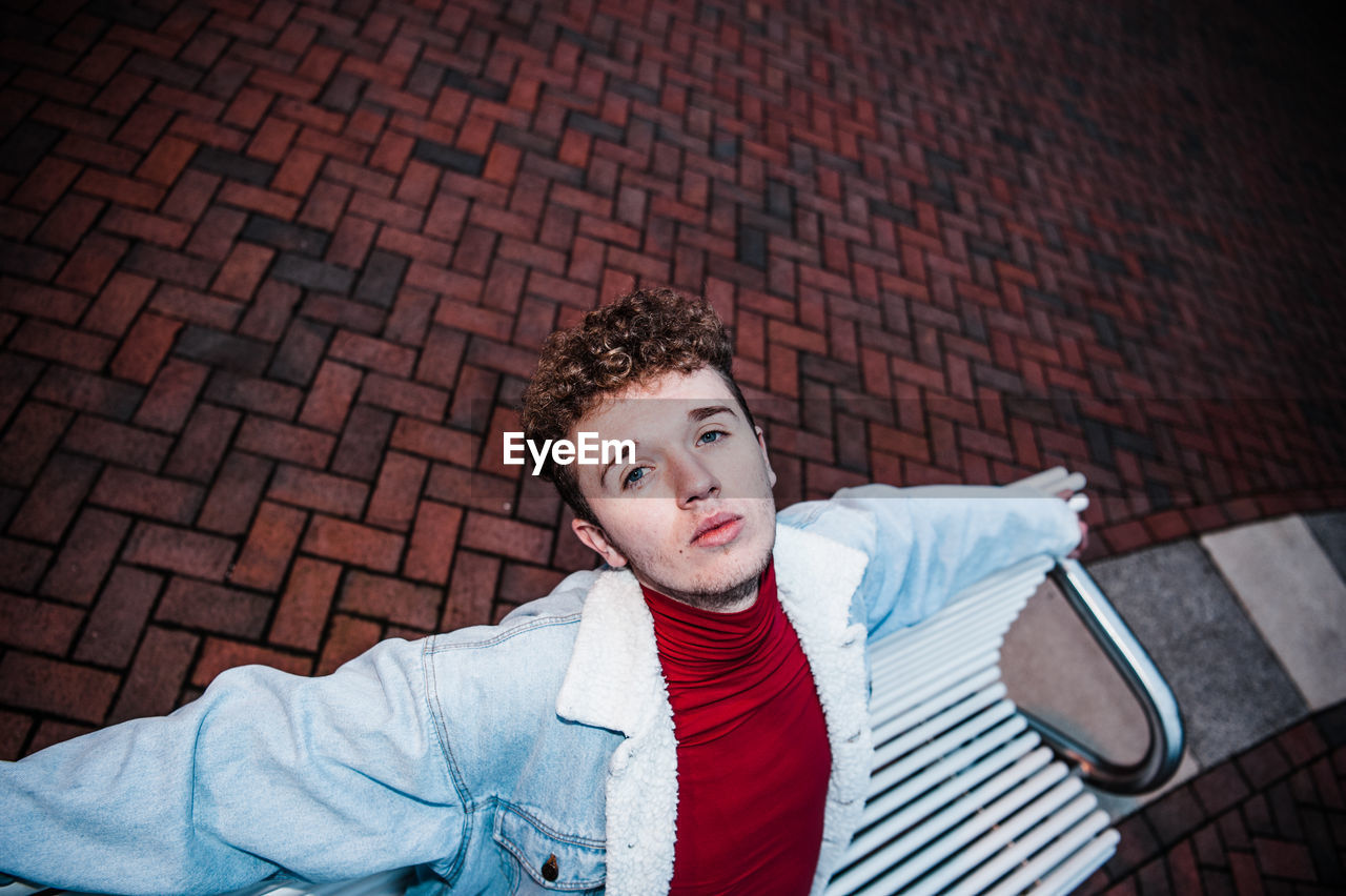 PORTRAIT OF YOUNG MAN SITTING AGAINST BRICK WALL OUTDOORS