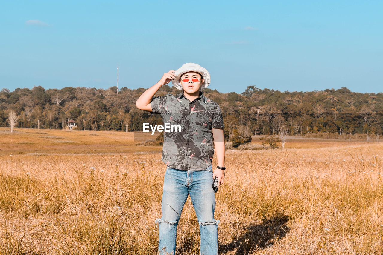 one person, landscape, field, sky, nature, plant, rural scene, land, adult, agriculture, front view, standing, environment, hat, casual clothing, portrait, copy space, day, full length, clothing, emotion, young adult, blue, women, grass, rural area, outdoors, jeans, looking at camera, smiling, prairie, crop, sunlight, farm, happiness, leisure activity, scenics - nature, lifestyles, sunny, fashion, person