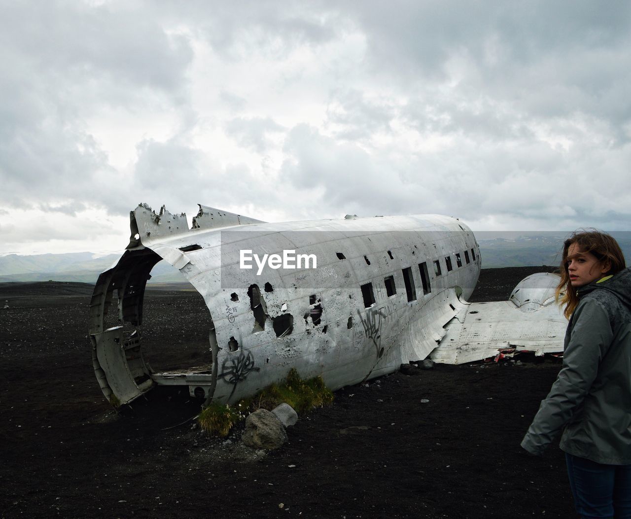 Young woman standing by abandoned airplane on field against cloudy sky