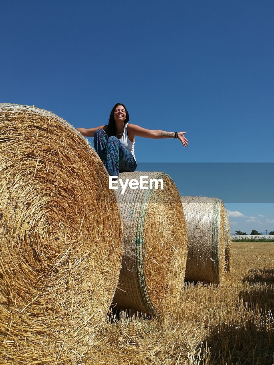 Low angle view of cheerful young woman sitting on hay bale against clear sky