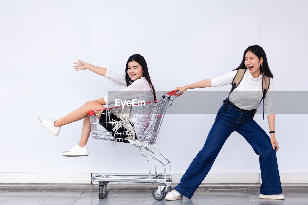Smiling young woman pulling friend sitting in shopping cart on tiled floor
