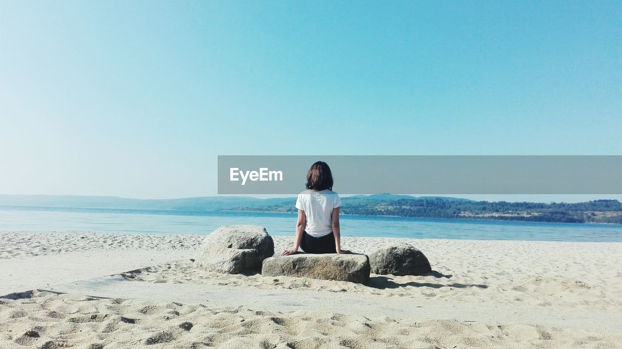 Rear view of woman sitting on beach against clear sky