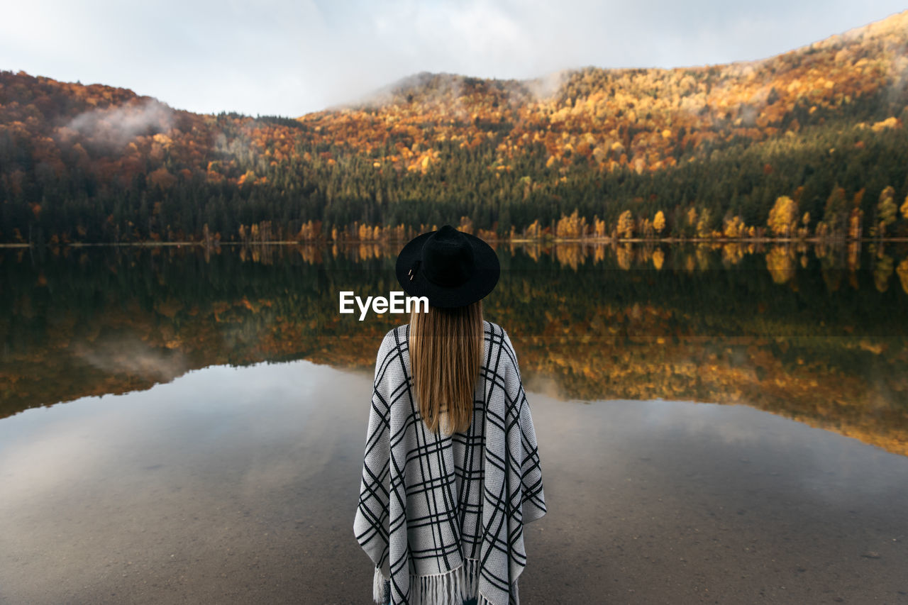 Caucasian woman standing in a poncho and hat. autumn in colorful forest near the lake.travel concept