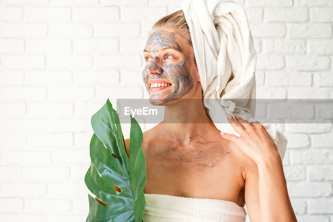 Smiling woman wearing bath towels with a clay facial mask on her face holding a green monstera leaf