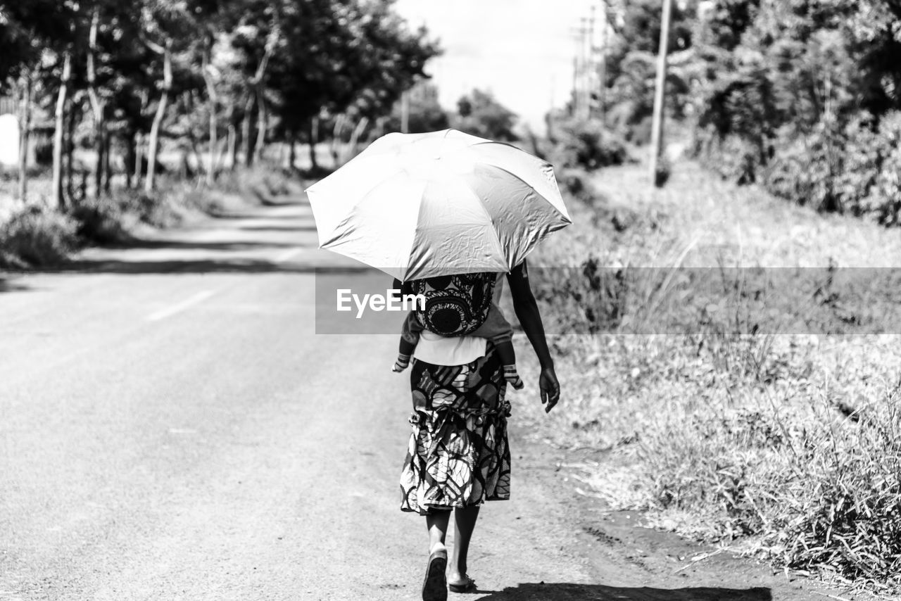 Rear view of woman with umbrella carrying baby while walking on street during sunny day