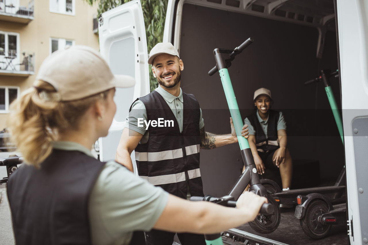 Smiling male worker talking with female coworker while loading push scooter in delivery van