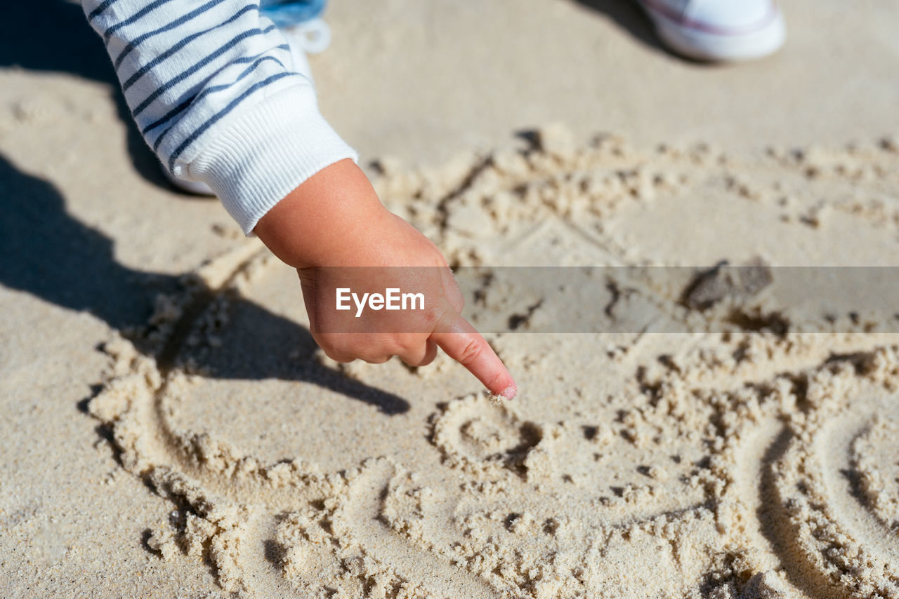 From above crop hand of little child in striped shirt drawing with finger on wet sand of coastline in sunlight